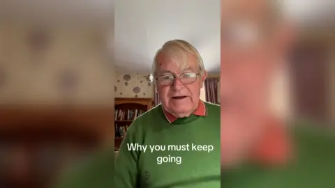 Ian Smith, and 81-year-old, dressed in a bright green jumper with orange polo shirt underneath. He has short white hair and is wearing thin black framed glasses.