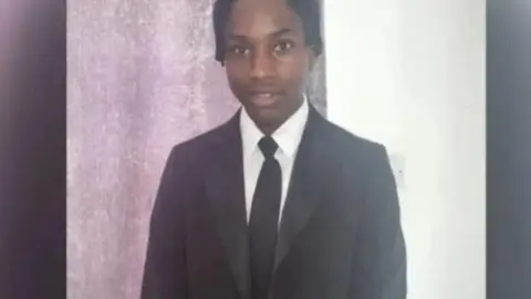 GOFUNDME Ashraf Habimana: A boy with black hair, wearing a black jacket, white shirt and black tie. 