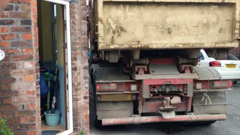 A lorry has mounted the pavement on the corner of a road. It appears to be very close to a house, which has an open front door.