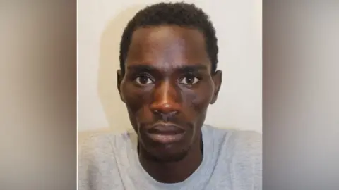 Marwan Jumaa has black curly hair and a grey prison sweatshirt and looks directly at the camera in what appears to be a police mugshot.