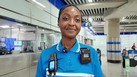 A station staff member in a blue uniform stands near the ticket gates in a bright, modern station. A radio and body-worn camera are attached to the uniform, with a control room visible behind glass windows and accessible ticket barriers in the background.