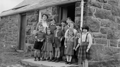 National Library of Wales Black and white photograph showing very young school pupils outside the school of Capel Celyn. Ten young school pupils can be seen huddled on the school steps. The boys were shorts, jumpers and shirts. While the girls were tartan skirts and cardigans. Their teacher stands tall at the back of the group, they all smile at the camera. 