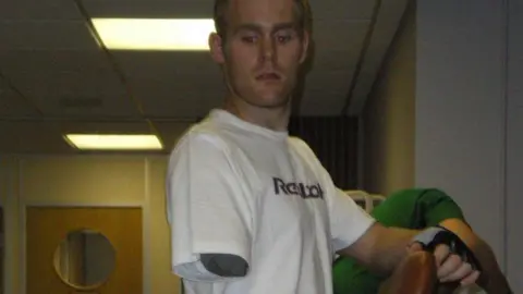 Mark Ormrod Mark Omrod with prosthetic arm and leg standing beside exercise or medical equipment in an indoor room with ceiling lights.
