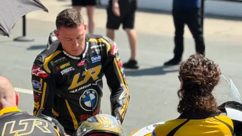 Channing Tatum is wearing black and gold leathers and is leaning over slightly, reaching for a matching helmet, with people stood around him, on a sunny day.
