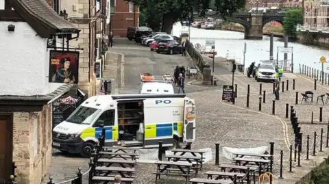 Picture shows a pub on the left with wooden tables and seats on a cobbled area outside the pub. Behind the tables and seat is a police van with it's side door open and towards the top of the picture is the river with another police car in the background.