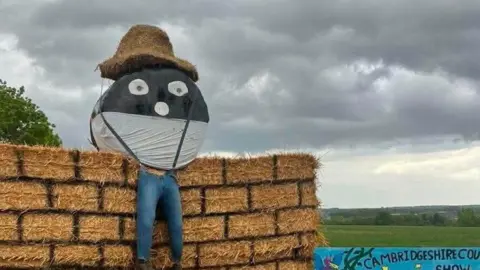 Huntingdon YFC A block of straw bales, with painted lines to look like bricks and perched on top a black and white round plastic coloured bale, with eyes and a nose with its legs made from stuffed blue jeans dangling below. To the side of the straw bale is a sign saying Cambridgeshire County Show 30/05/26 and beyond it green fields under grey skies.