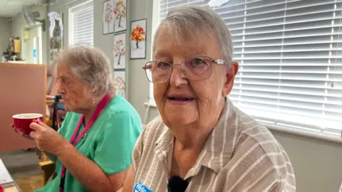 Renee is wearing glasses and a tan top with white stripes. She has short hair and a lady is next to her drinking a cup of tea in a red cup. 