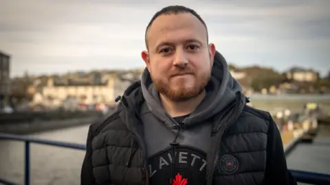 Un hombre con cabello corto y oscuro y una barba de color claro mira directamente a la cámara. Está parado en Bute Harbour, con agua, un muelle y casas en el fondo. Lleva un saltador con capucha gris y un calentador de cuerpo negro.