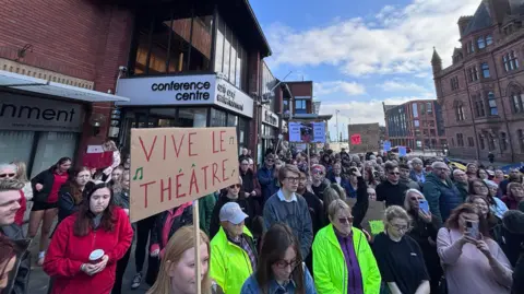 Shot of dozens of people standing outside in the centre of a market town on a sunny day. In the background is a building which says Conference centre - its a grand old brown brick building. One of the people has a cardboard sign which says Vive Le Theatre.