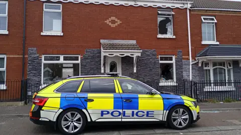 A police car is parked outside a red and grey brick terrace house. 