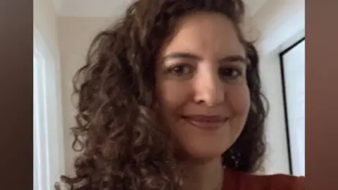Dr Elizabeth Nassem A woman with long curly brown hair wearing an orange jumper