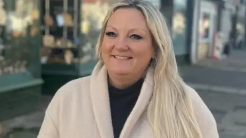 Portsmouth Labour Party Amanda Martin MP - with long blonde hair, wearing a cream coast smiling and looking at the camera. Shops can be seen behind her.