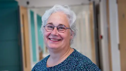 Bradford Teaching Hospitals NHS Foundation Trust A white-haired woman wearing glasses and a blue top with green and white petal details. 
