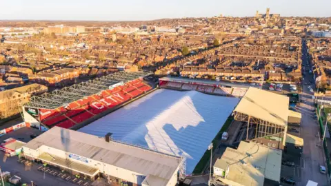 Chris Vaughan/LCFC Aerial view of a football stadium with a protective cover, resembling a huge white tent, over the playing surface. There are four stands surrounding the pitch. The word "city" can be seen on one of the stands, which has red seats, and there are houses and a large cathedral in the background.