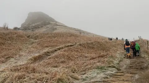 Lottie King Roseberry Topping which is a large hill with frost-covered grass leading up to its peak. People can be seen walking up the stairs towards it.