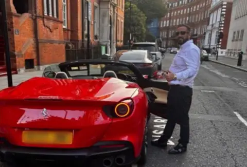 Facebook A man with dark hair, a stubble beard and sunglasses is standing beside a red Ferarri car, which has the roof lowered and the door open. He is dressed in smart black shoes with black trousers and a blue shirt, and is wearing a gold watch. The man is giving a thumbs up and is looking at the camera. The car is parked on a street with other vehicles and large red bricked buildings on either side. 