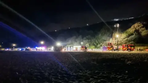 DWFRS It's nighttime along Bournemouth seafront, there are fire engines lining the promenade and spotlights and some smoke on the hilltop