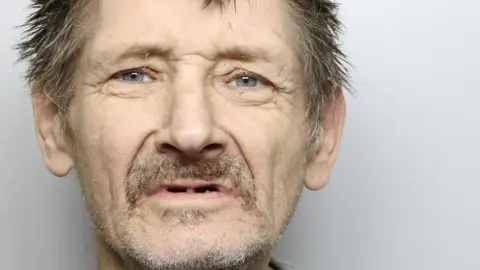 A custody picture of a man with messy grey hair and facial stubble. 