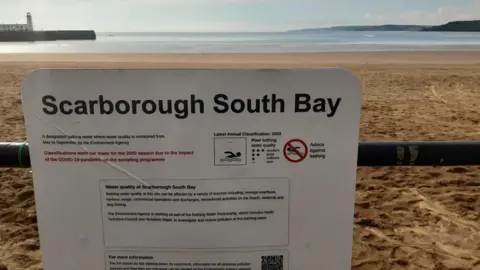 Anttoni James Numminen A sign affixed to a metal barrier at Scarborough's South Bay warning of the bay's bathing water status. In the background, sand, sea and a lighthouse can be seen.