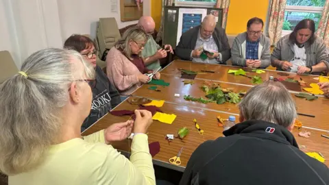 Cad Taylor, BBC Numerous people are sitting around a table making things. The table is littered with scissors, pieces of coloured felt and leaves