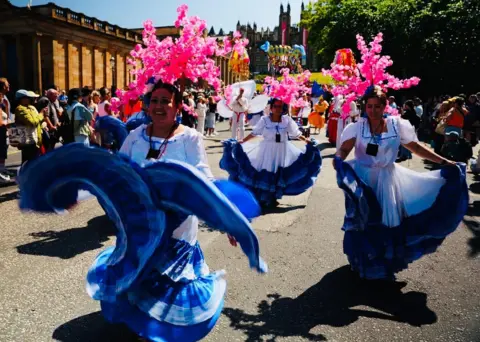 A group of parade performers in white dresses with blue skirts dance along a city street decorated with large pink blossom headpieces. Crowds watch from both sides of the parade route on a sunny day.