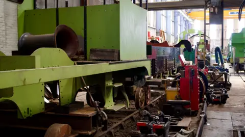 An engineering shed at a train depot. A light green locomotive frame is on a track surrounded by engineering equipment.