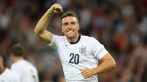 Getty Images Former Bristol Rovers player Rickie Lambert punches the air as he celebrates a goal for England against Scotland. He is wearing the traditional all-white England shirt
