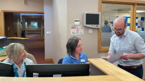 Mark Ansell/BBC A fair-haired woman in a nurse's uniform and a dark-haired woman in a nurse's uniform sit behind two computers at a counter. A man with glasses and facial hair and wearing a shirt and navy trousers is showing them a folder.