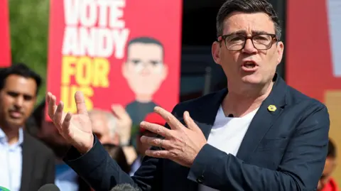 Reuters Andy Burnham, 56, who has black hair with streaks of grey and black framed glasses, speaks to a crowd while wearing a dark blazer over a white t-shirt. In the background supporters hold red placards with the slogan 'Vote Andy For Us'. 