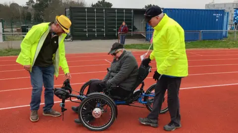 Two men in fluorescent jackets help an older man onto a three-wheeled adapted bicycle. 