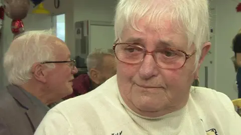 Janet is wearing a white Christmas jumper and has white hair with glasses on.