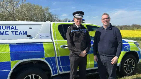 PCC John Campion wearing a navy blue quarter zip jumper is stood beside Chief constable Richard Cooper, wearing a black police uniform. The pair are stood in a field in front of a large 4x4 police vehicle which has been painted yellow, blue and white.