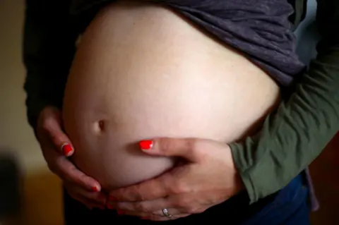 PA Media A pregnant woman is holding her bare stomach. She is wearing a blue and green top and blue jeans. She has red nail paint on.
