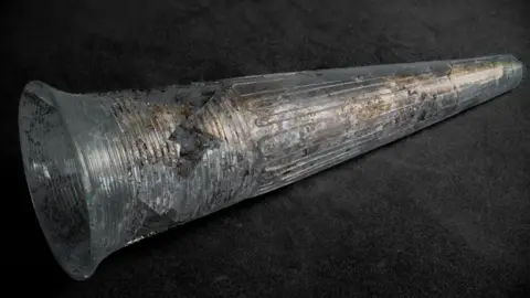 Discover Bucks Museum A glass conical drinking beaker lying on its side with its wide rim on the left narrowing to the right. It is decorated in fine evenly spaced lines of flace. It still has mud clinging to it. Found at Wendover.