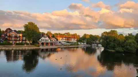 AliB The River Thames reflects the fluffy clouds and blue and yellow skies next to a boathouse and homes on it's banks. A few birds can be seen floating on the water.