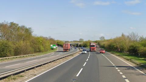 Cars and two lorries drive on a dual carriageway on a major road, with trees and green foliage on either side. 