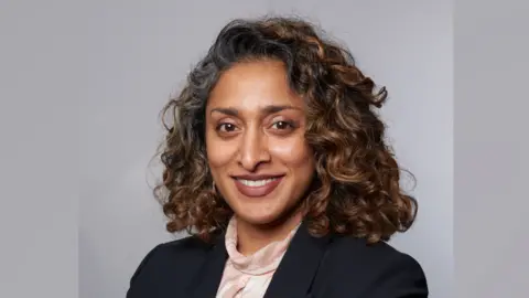 Essex & Suffolk Water A head and shoulders image of Monisha Gower. She is wearing a black blazer over a light top. She is standing in front of a grey backdrop and smiling while looking into the camera.