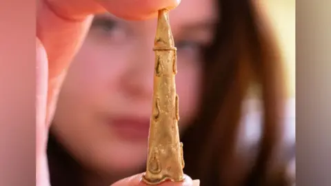 Lori Rogerson Lori Rogerson holding up a Roman gold conical earring. The image is focused on the earring, which she is holding in thumb and forefinger, with the pointed end uppermost. She can be seen in the background, with dark hair, but out of focus