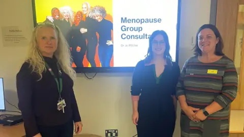 Three women are standing in front of a large television screen that has a photo of women on it and reads "menopause group consultation". The woman on the left has blonde hair, a black top and a green lanyard. The woman in the middle has brown hair, glasses and a black and green top. The woman on the right has short brown hair and a grey, black and red striped top with a yellow name badge.