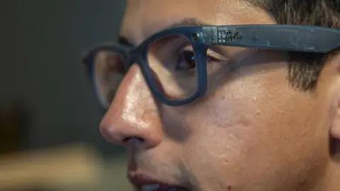 Getty Images Close-up profile of a person wearing translucent, dark grey Ray-Ban smart glasses featuring a small built-in camera lens near the hinge.