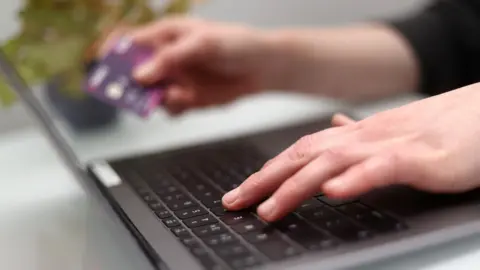 PA Media A woman using a laptop as she holds a bank card