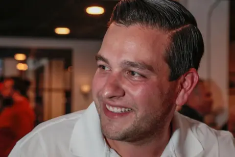 Getty Images A man in a white shirt smiles, with his face pointed away from the camera