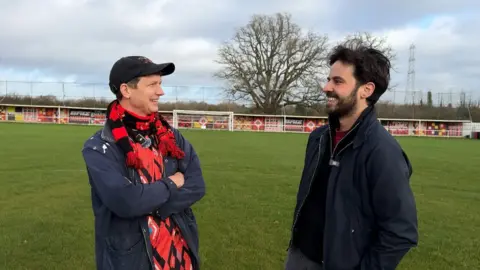 John Kelham and Ken Borg smiling at each other on the pitch.