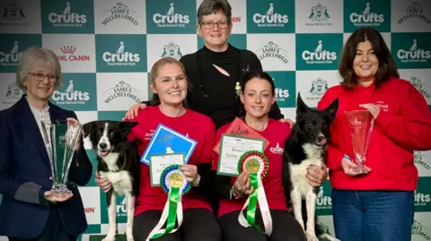 Sam Springford Three ladies in red tops, with black and white dogs either side under the arms of two women. They are in front of a green and white banner, with another lady in a blue blazer holding a trophy on the left and a woman in a black top behind them.