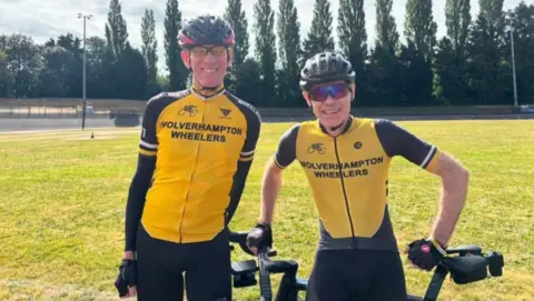 Two men wearing mainly yellow tops including the words Wolverhampton Wheelers. They are wearing glasses and cycling helmets and a large area of grass and trees are in the background.