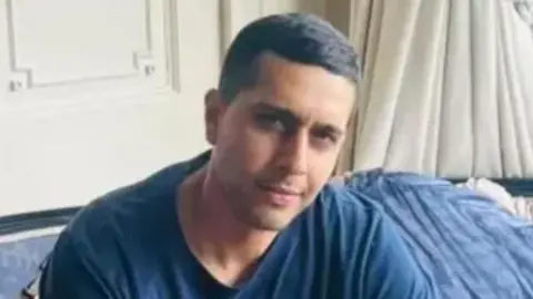 Kamran Aman sitting on a blue sofa in a living room with white walls and white curtains visible behind. He wears a navy blue t-shirt and has short black hair