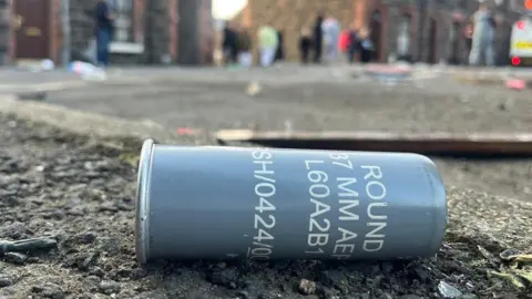 A close up image of a large metal tube on the ground of a street - there are words and numbers printed on the grey casing, one of which is 'round'. In the background people can be seen standing a distance away