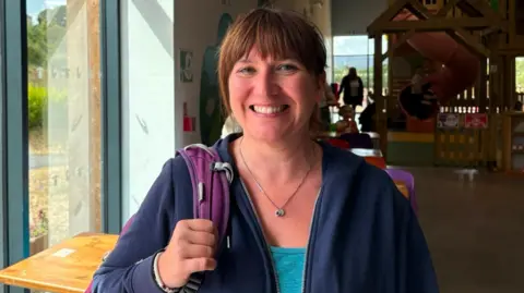 A woman wearing a rucksack, a blue unzipped hoodie and a blue top. She is stood in front of a play centre for children, slide viewable in the background.