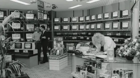Metrocentre A black and white photo of the technology shop Currys in the 80s. There is a blonde woman with a perm bent over behind the counter. Two men talk in the left corner of the shop. The back wall is lined with old, small televisions. There is a plinth with Currys written on it in the centre of the photo.