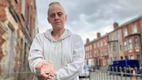 A woman is standing in a small urban garden area with a long street behind her. It is lined with red brick houses and cars parked along the pavement. The woman has blonde hair tied up in a bun and is wearing a grey jumper. She looks directly at the camera while holding two loose keys in her hand. 